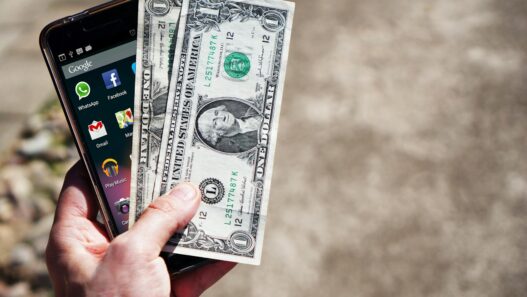 A person checking their checking account balance on a banking app, with cash and a notebook nearby
