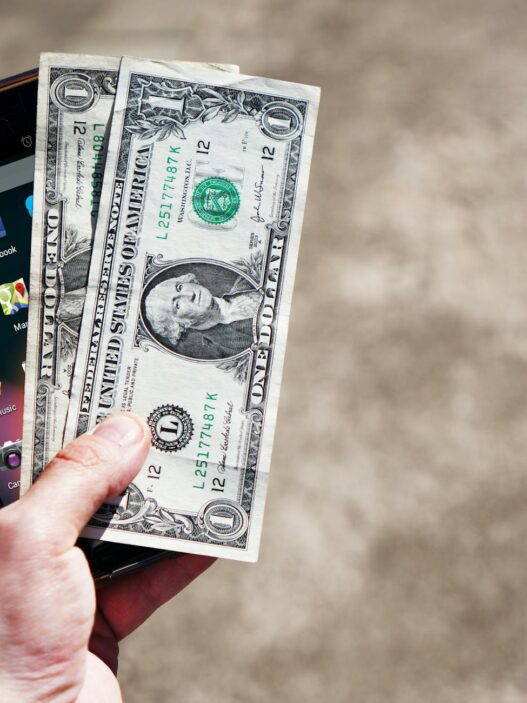 A person checking their checking account balance on a banking app, with cash and a notebook nearby