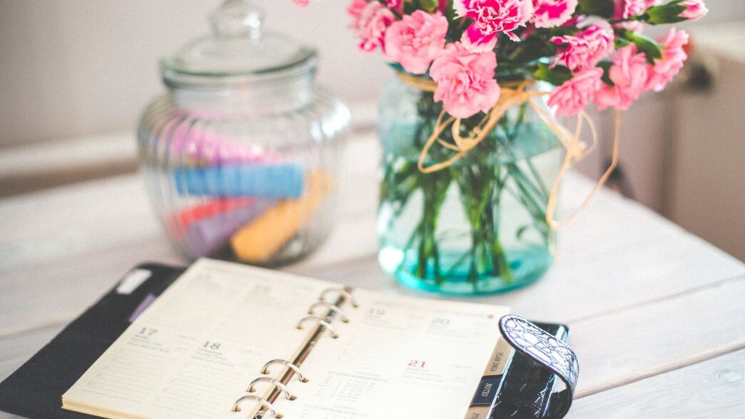 A monthly calendar and notebook beside a jar of coins on a desk